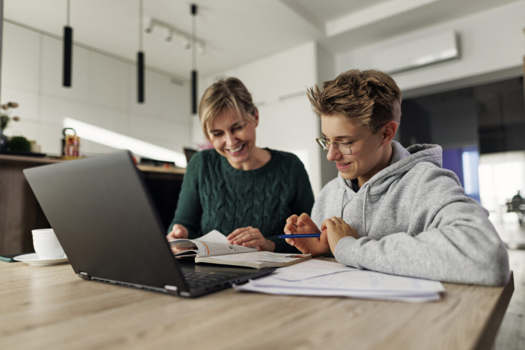 A woman and a boy sit at a table with a laptop, books, and papers, smiling and studying together in a modern kitchen.